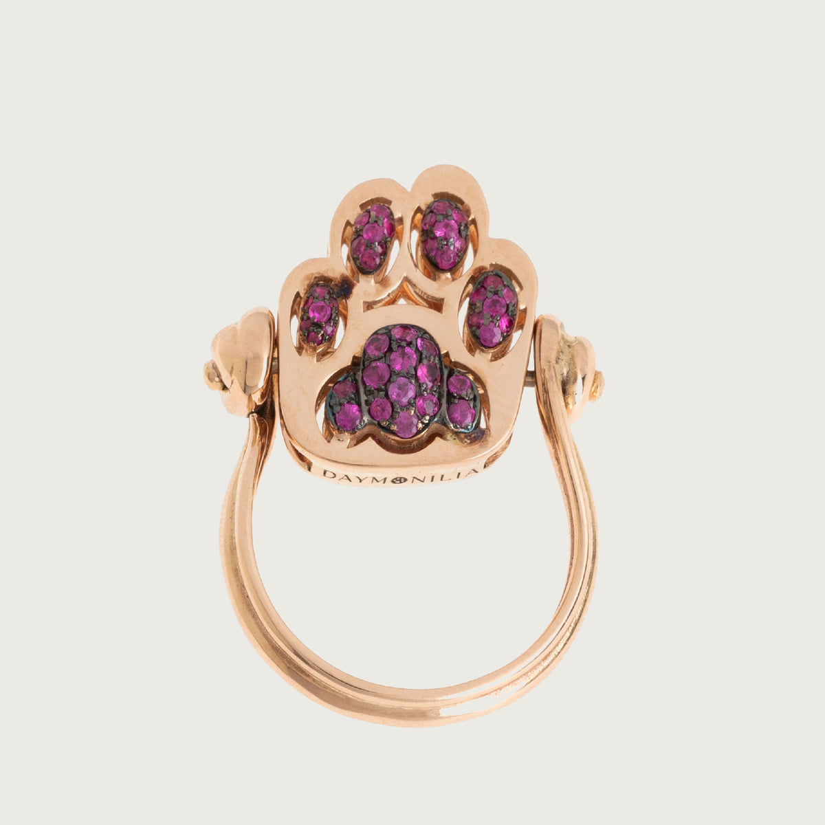 Fusa: anello Daymonilia in oro rosa con la zampa di rubini, visto di fronte su fondo osso.